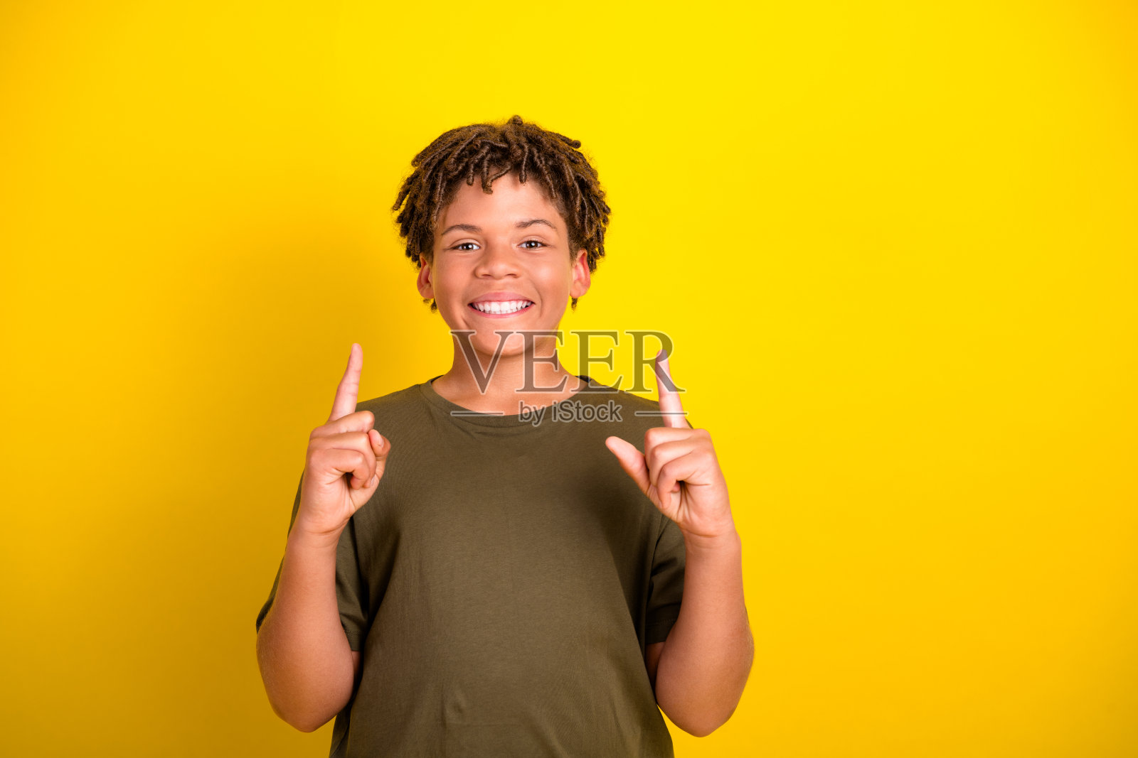 一个穿着卡其色T恤的、不同族裔的男孩,脸上洋溢着笑容,向上指着,背景是明亮的黄色,营造出欢乐的生活方式推广氛围照片摄影图片