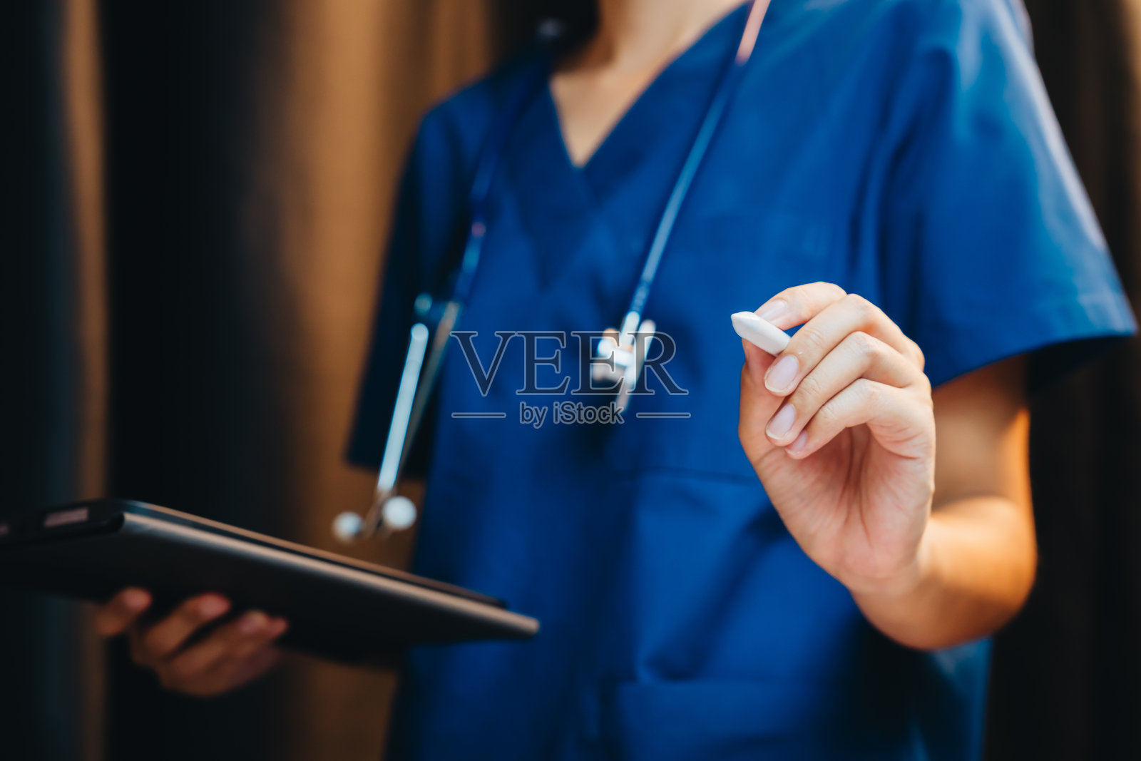 身穿蓝色手术服的医护人员手持平板电脑和触控笔,听诊器挂在脖子上,身处临床环境照片摄影图片