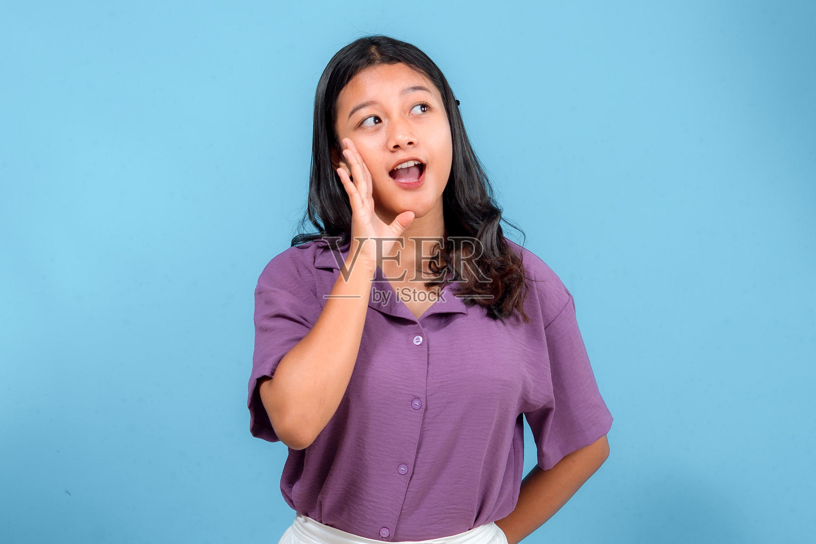 年轻亚洲女性表情丰富,手靠近嘴边大声呼喊或叫喊,展现出兴奋和惊讶的情绪。非常适合广告、沟通和公告主题。照片摄影图片