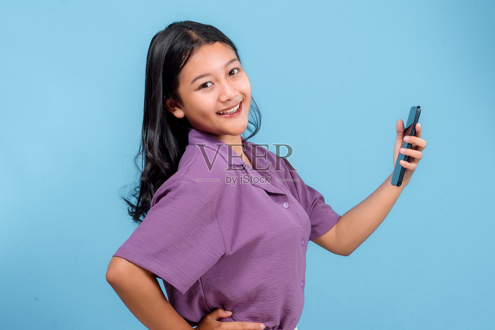 一位面带快乐自信表情的年轻亚洲女性,手持智能手机,站在蓝色背景前。非常适合数字营销和科技广告。照片摄影图片