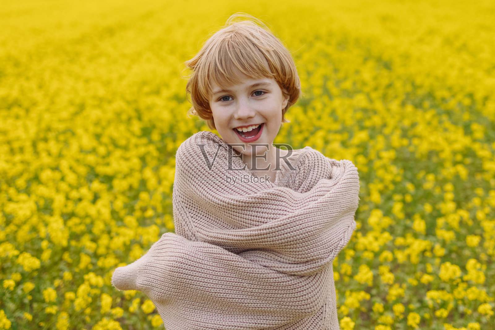 穿着宽松米色毛衣的女孩在黄色花田中拥抱自己,面带微笑照片摄影图片