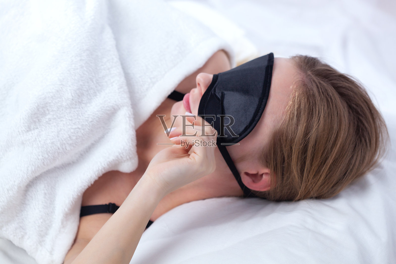 一位年轻美丽的棕色头发女孩戴着口罩,在早晨的白色床上睡觉。照片摄影图片