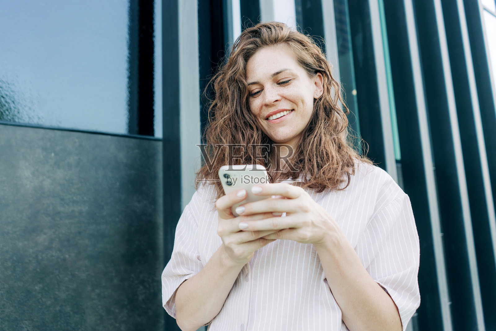 一位美丽的年轻高加索女性,留着波浪卷发,在户外建筑旁使用手机。照片摄影图片