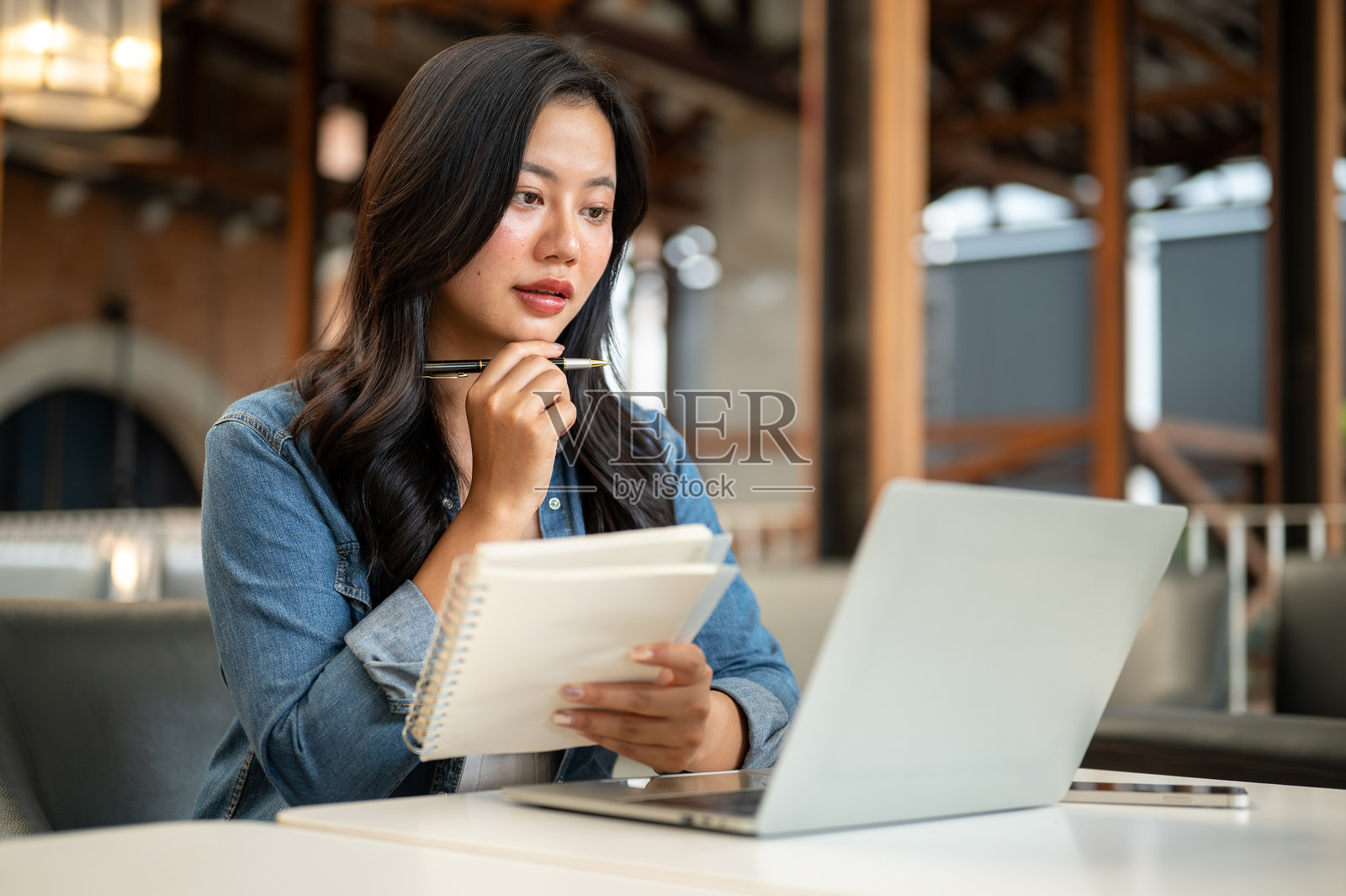 一位微笑着的亚洲女学生,下巴托着笔和笔记本,看着咖啡桌上的笔记本电脑。照片摄影图片