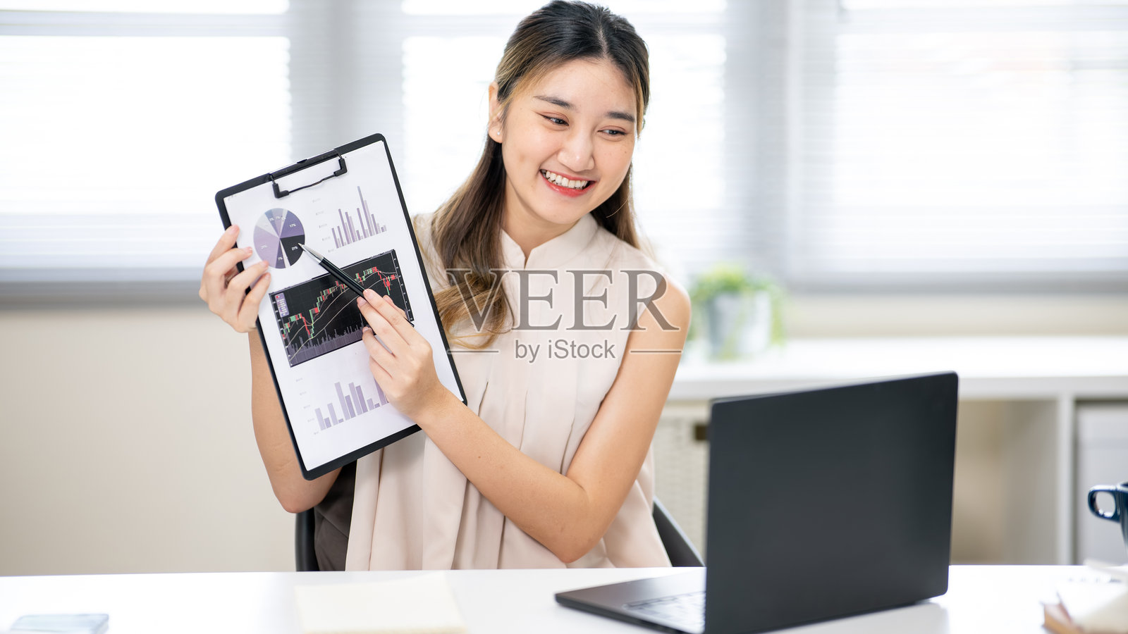 一位漂亮的办公室女职员,手持钢笔,在办公桌前的笔记本电脑旁的剪贴板上指示。照片摄影图片