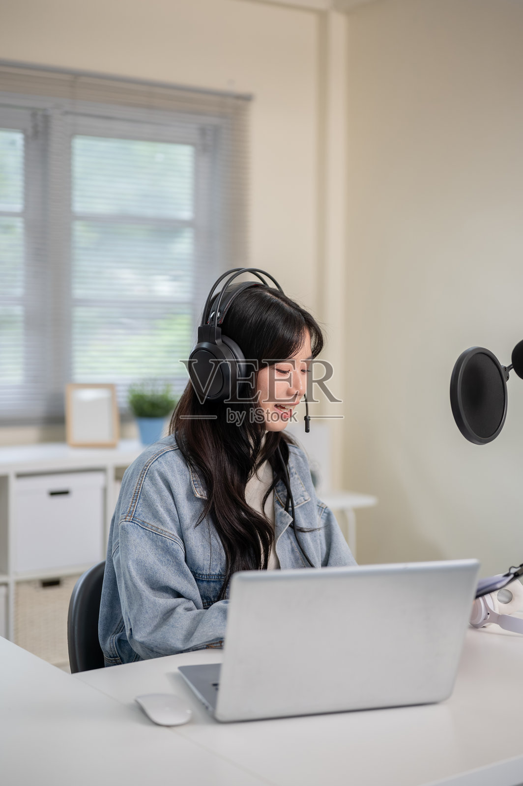 亚洲女性戴着耳机和麦克风,通过笔记本电脑主持广播或播客,同时查看剪贴板。照片摄影图片