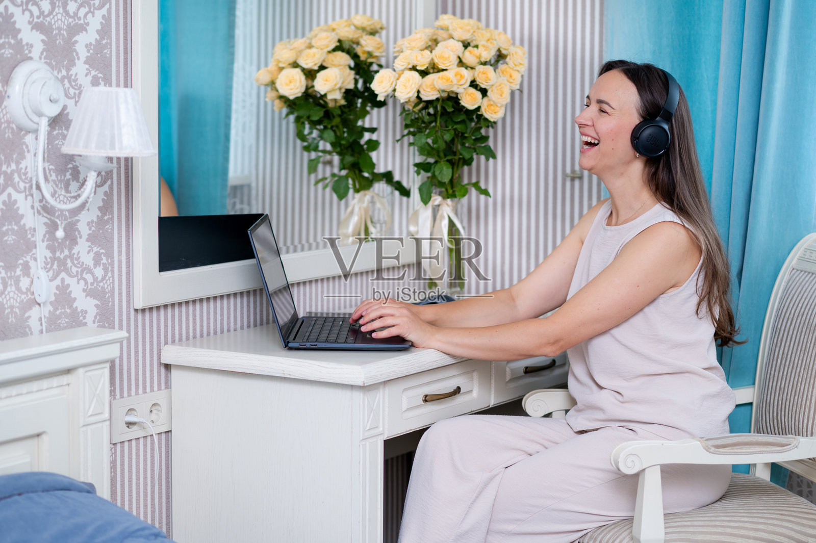 年轻女子戴着耳机在卧室里通过笔记本电脑进行交流,旁边放着一束黄玫瑰。照片摄影图片