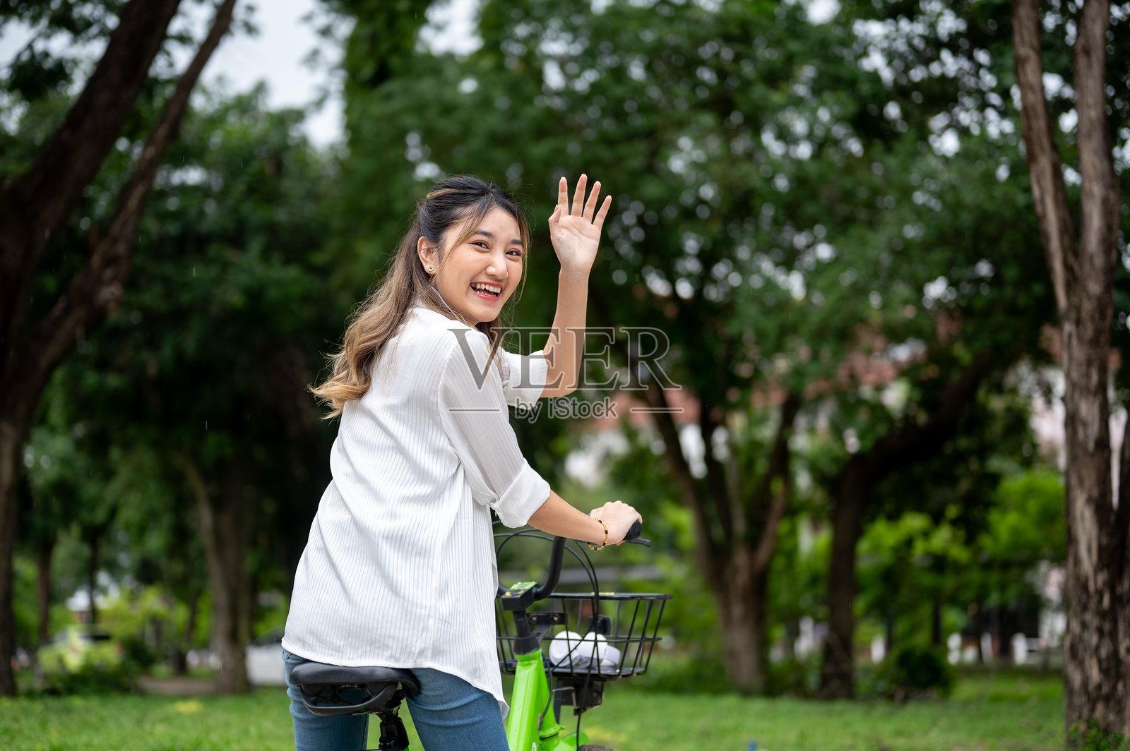 一位年轻的亚洲女性面带微笑,回头挥手,同时在自然公园里骑着自行车。照片摄影图片