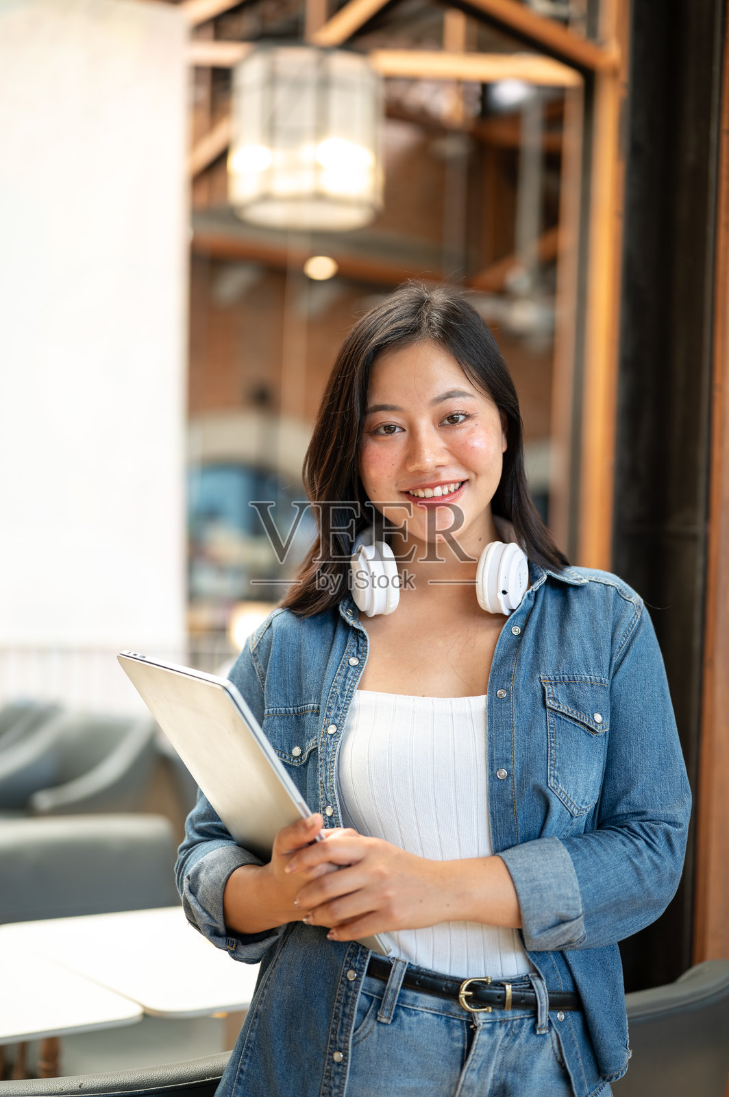 漂亮的亚洲女学生戴着耳机,手里拿着笔记本电脑,站在咖啡馆或咖啡店里。照片摄影图片