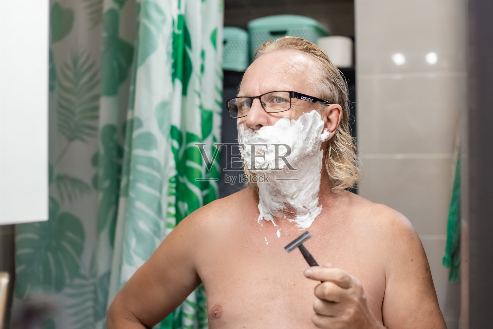 一位中年男子脸上涂着剃须泡沫,在浴室镜子前审视自己,准备刮胡子照片摄影图片