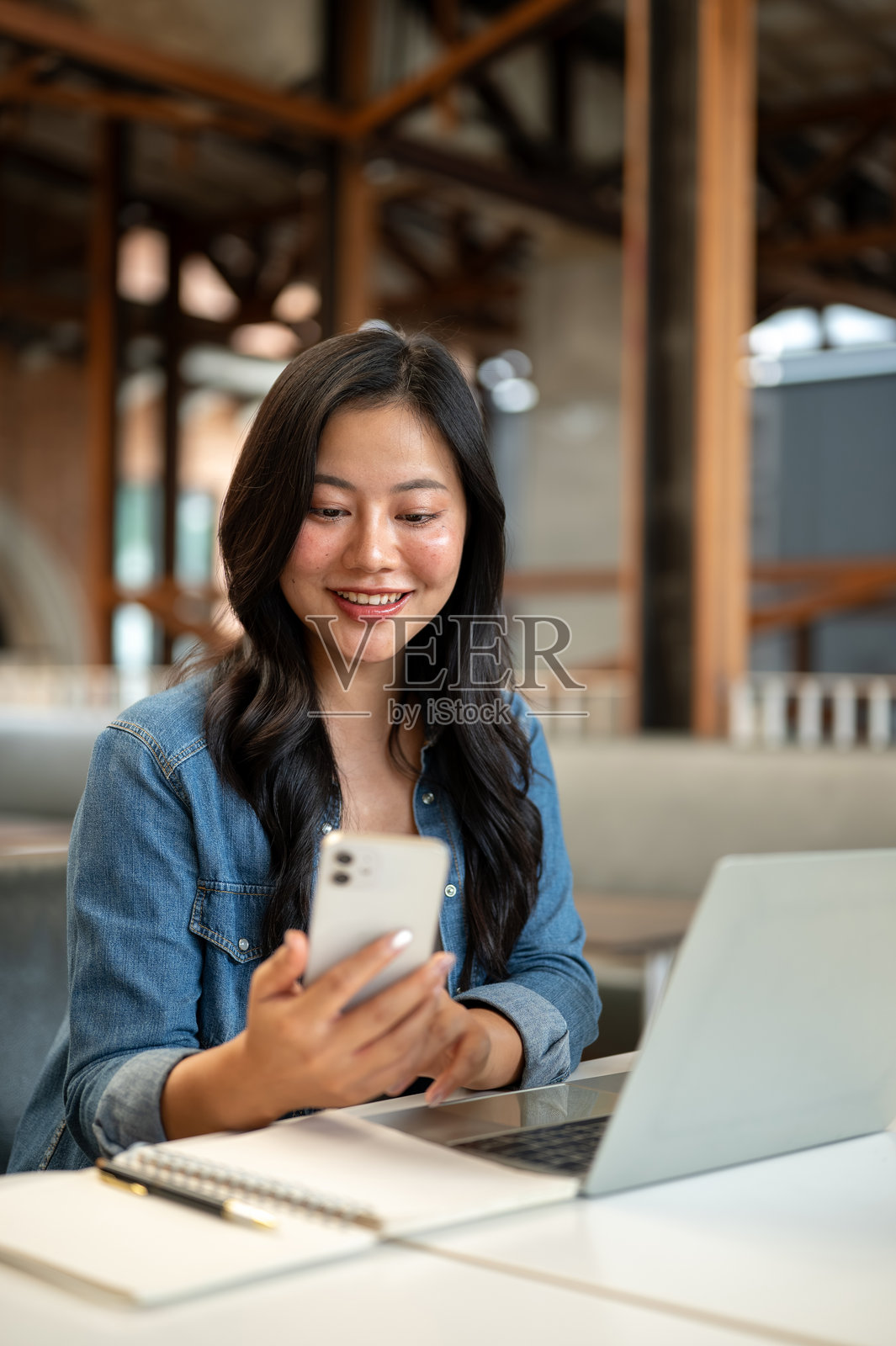 一位亚洲女学生坐在咖啡馆的桌子旁,笔记本电脑放在一旁,她手里拿着手机并看着手机照片摄影图片