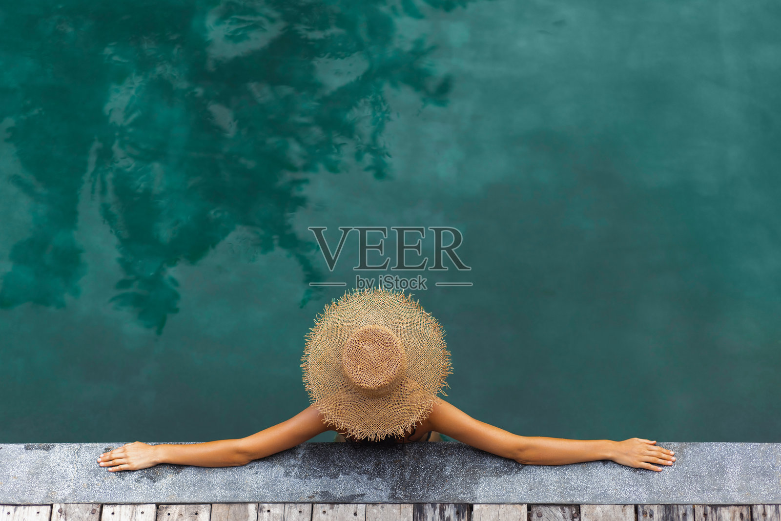 戴草帽的女子在泳池放松,手臂搭在木制甲板上照片摄影图片