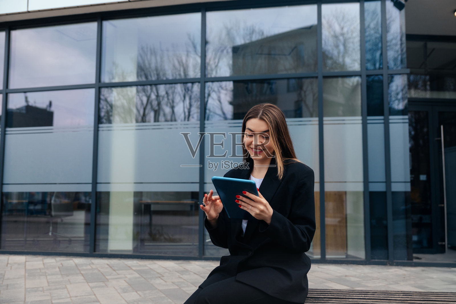 一位年轻女子在多云天气下,在现代化建筑外使用平板电脑,面带微笑,享受着时光。照片摄影图片
