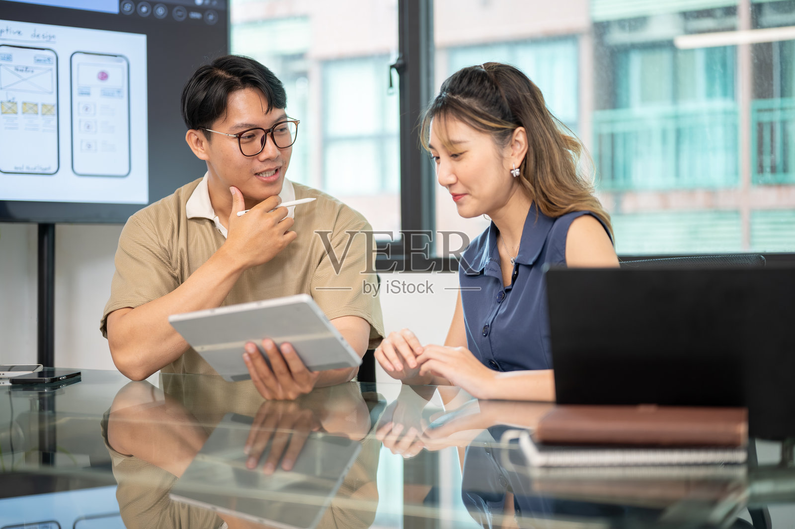 一位戴眼镜的亚洲男性在办公室会议室里,手持平板电脑和触控笔,与一位女性同事交谈。照片摄影图片