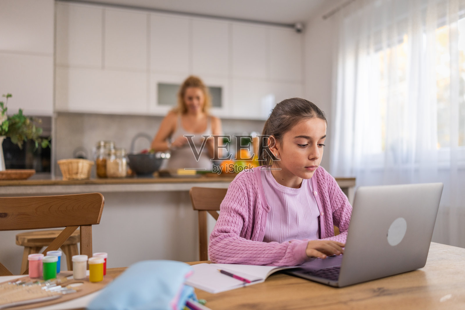 可爱的小女孩坐在餐桌前,在线做功课,背景中她怀孕的母亲在模糊中烹饪早餐。照片摄影图片