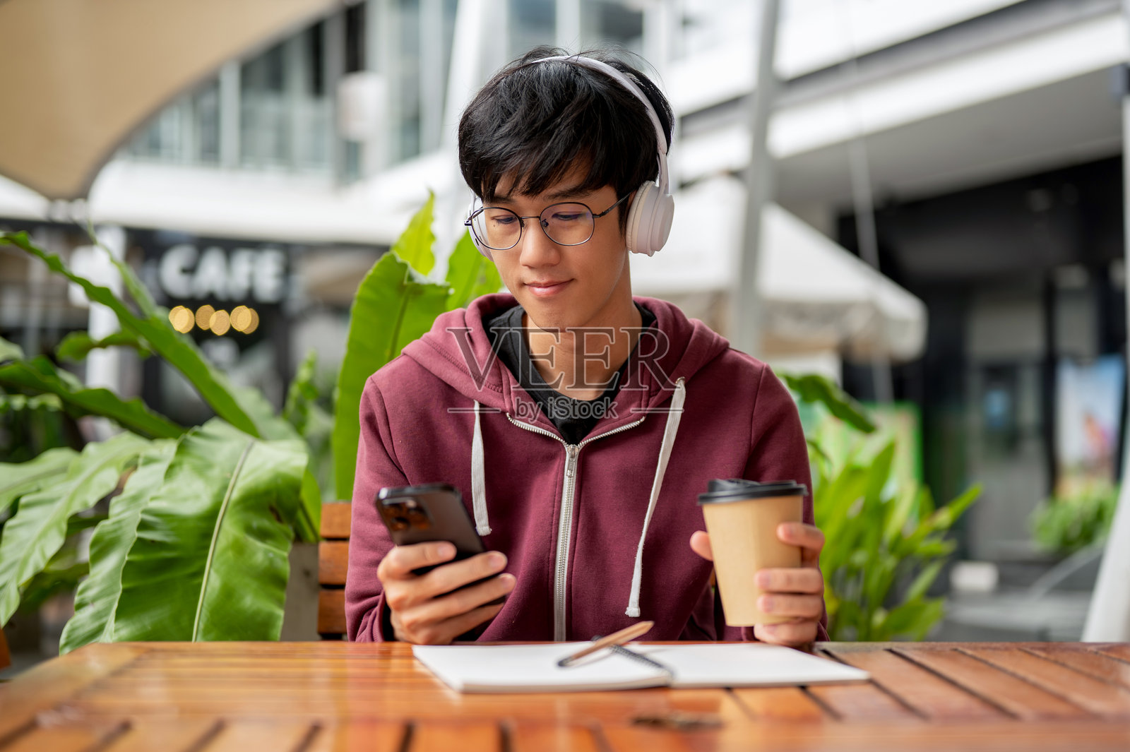 戴着耳机、拿着咖啡、看着手机的眼镜男学生坐在咖啡馆桌子旁,旁边放着笔记本。照片摄影图片