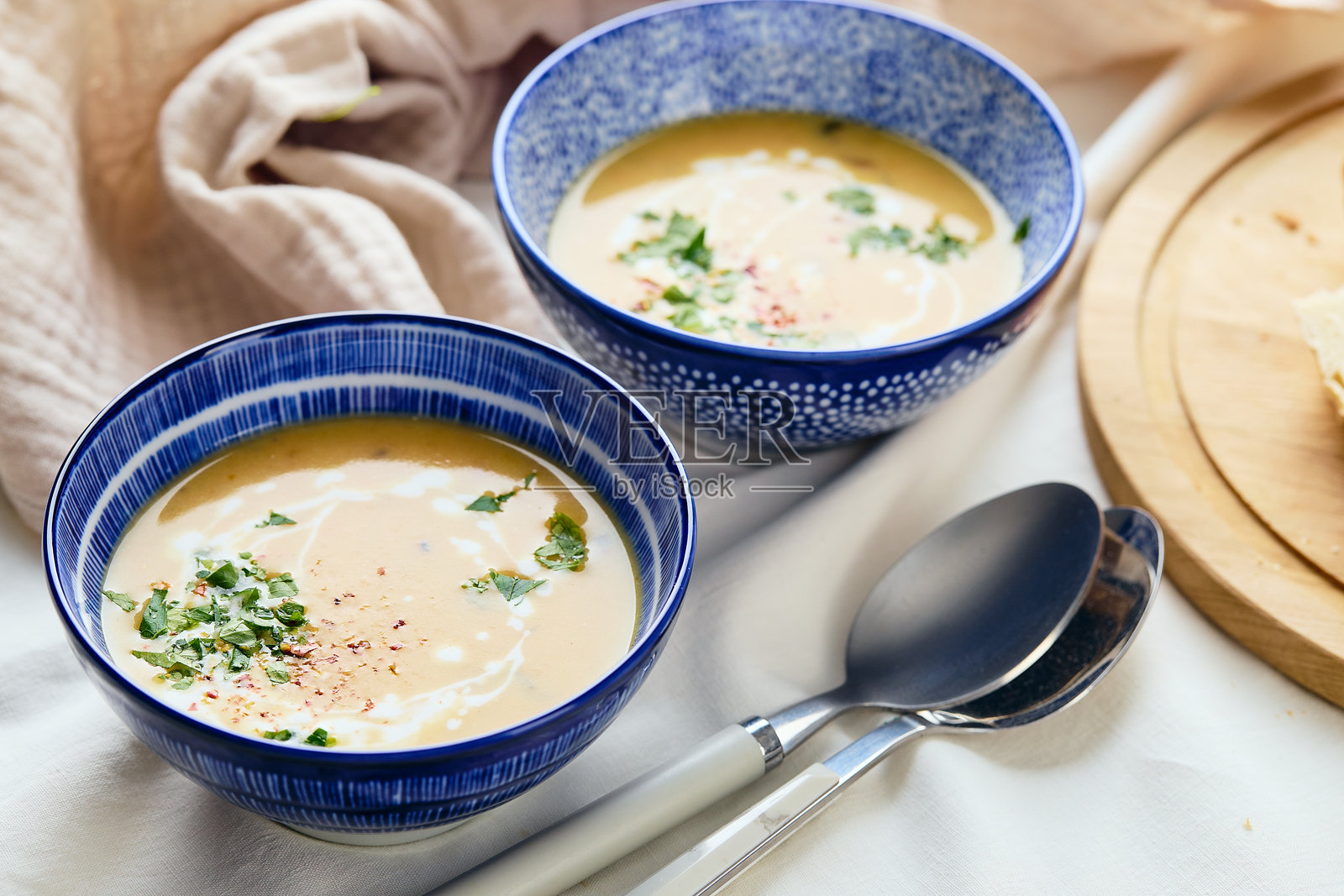 两碗奶油土豆浓汤,配有勺子、餐巾和木质砧板,砧板上放着一条面包和切好的面包块。汤的表面装饰有奶油、面包丁、欧芹和红胡椒粉。照片摄影图片