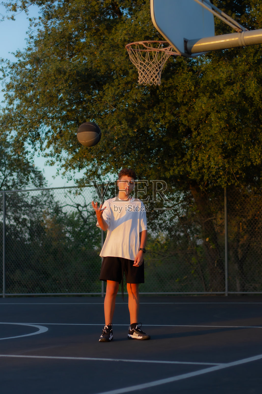 夕阳下,一个小男孩在郊区户外的篮球场上练习篮球,背景宁静祥和。照片摄影图片