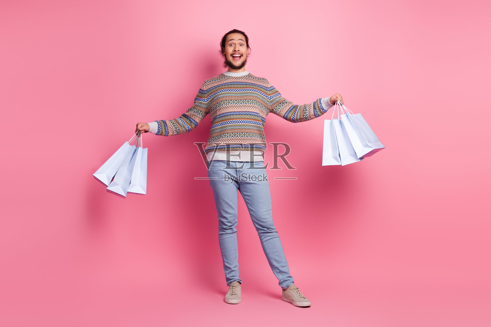 留着胡子的快乐年轻男子,身穿时尚休闲毛衣,拎着购物袋,背景为粉色。照片摄影图片