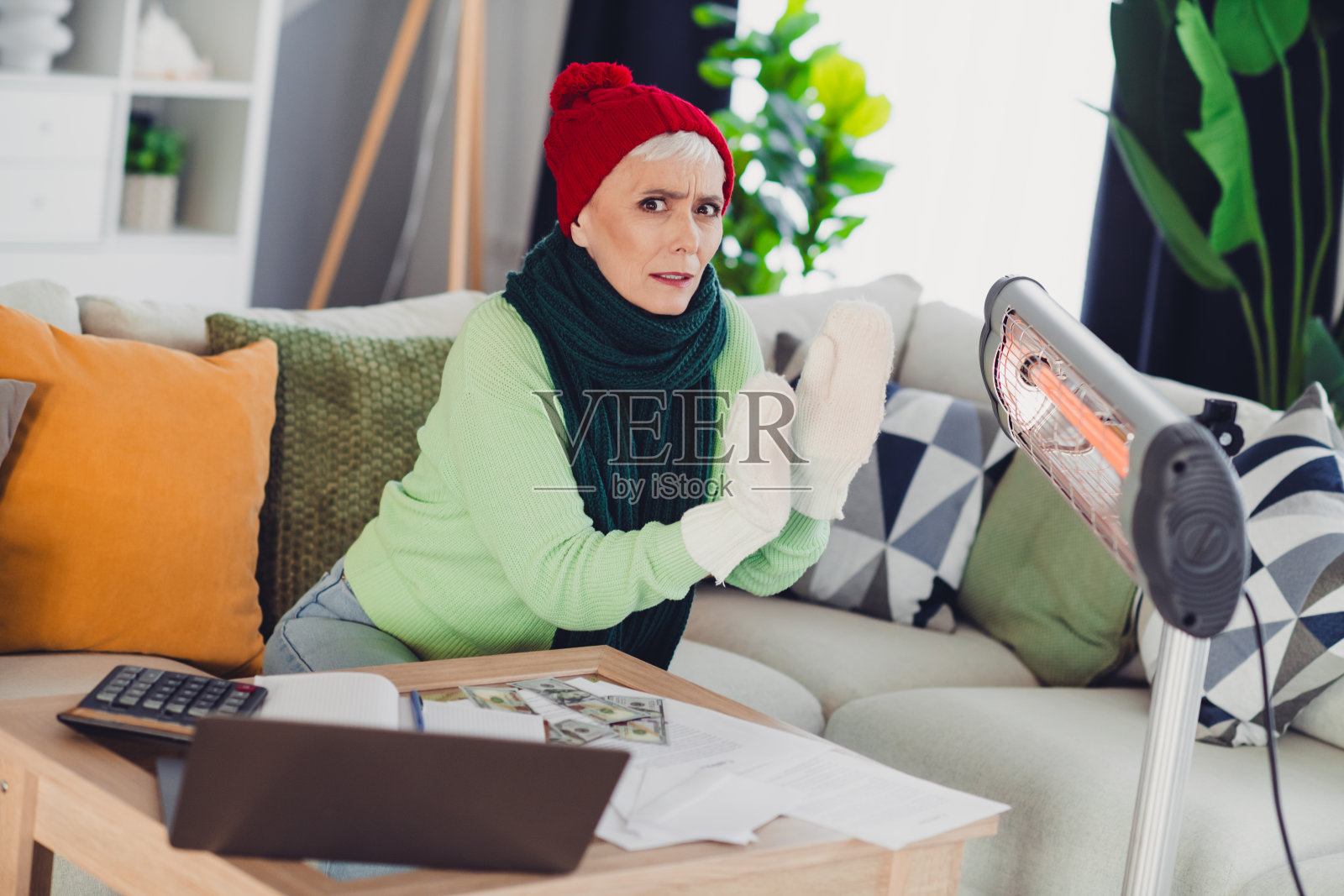 一位身穿舒适冬季服装的老年女性在客厅的暖炉旁暖手,周围散落着文件和一个笔记本电脑照片摄影图片