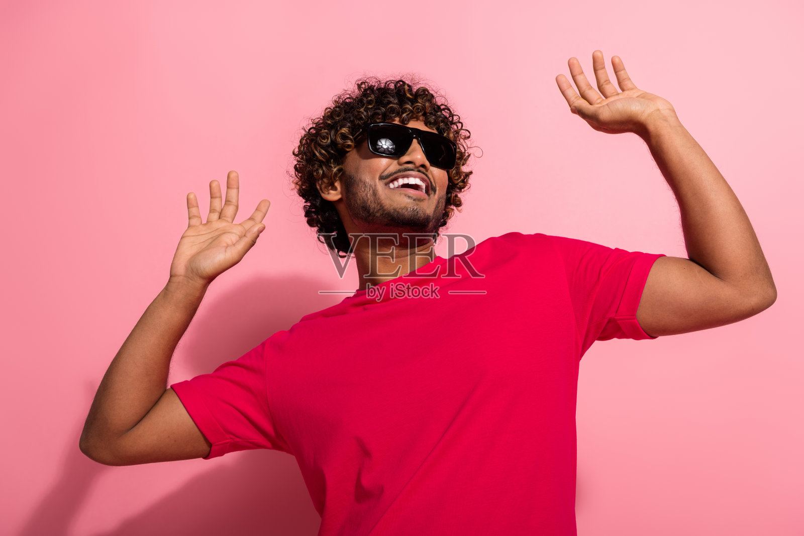 一个卷发年轻男子穿着鲜艳的红衬衫,带着快乐的手势,在粉色背景前摆姿势,拥抱积极乐观照片摄影图片