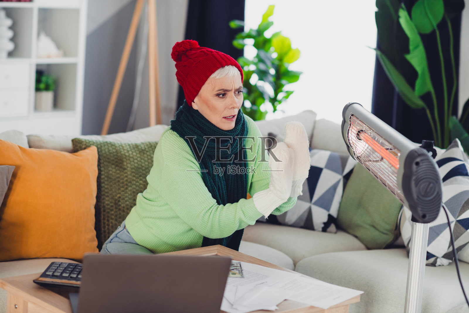 一位年长的女性在家中,身着舒适的冬装,靠着取暖器取暖,展现出室内的舒适与放松。照片摄影图片