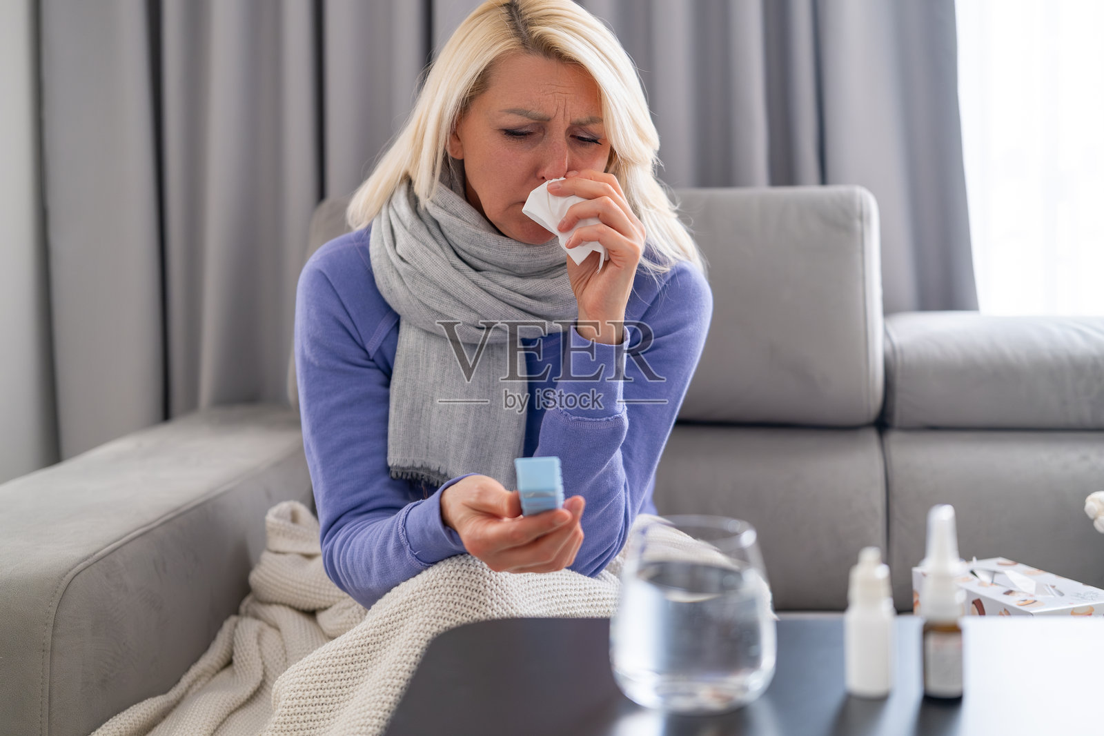 生病的金发女子在沙发上擤鼻涕,脖子上围着围巾,桌上放着药品和水照片摄影图片