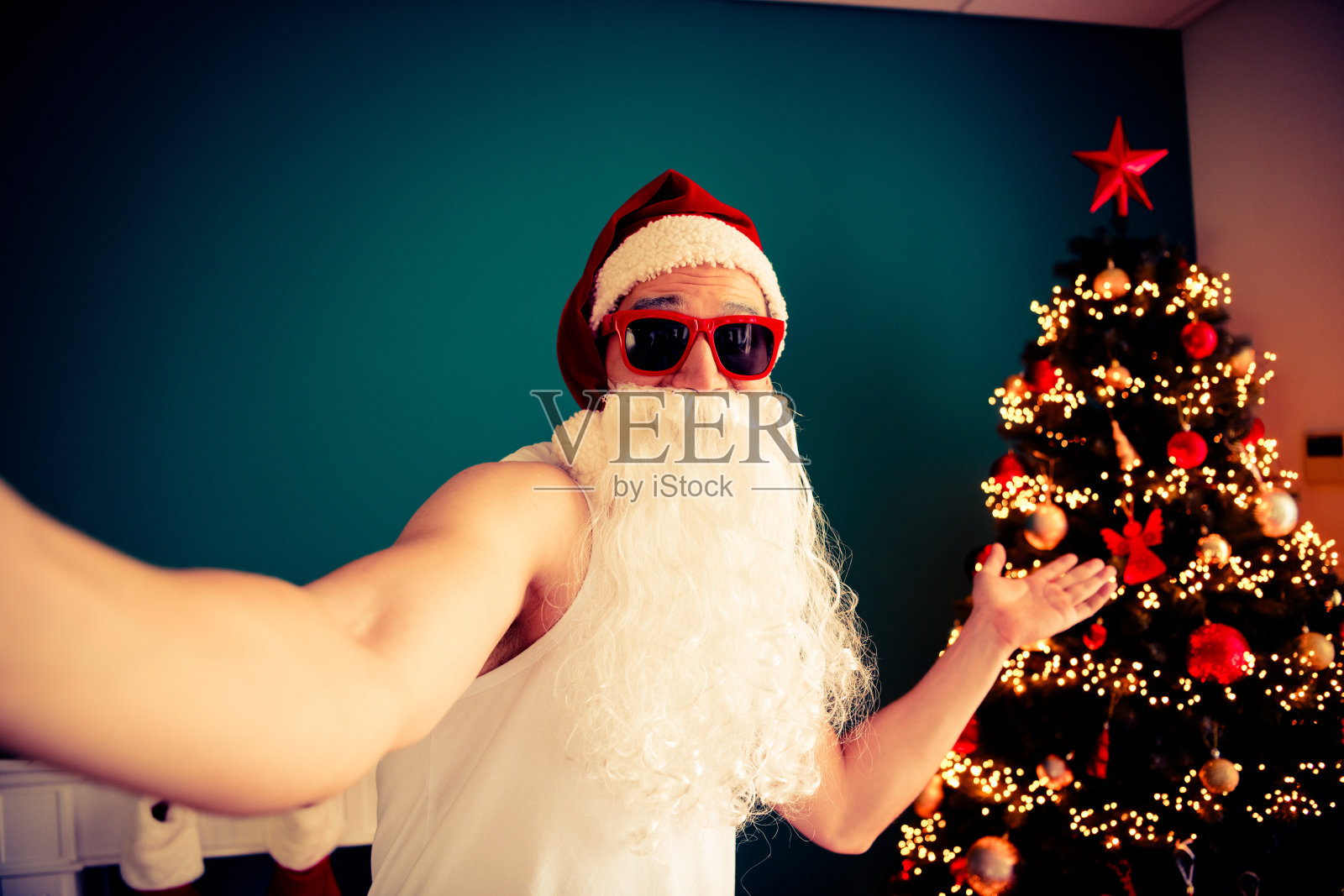 圣诞老人戴着太阳镜,在舒适的节日房间里,在一棵闪耀着节日灯光的漂亮圣诞树旁,拍了一张幽默的自拍照。照片摄影图片