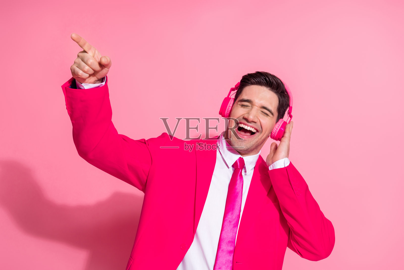 穿着粉色西装的年轻男子戴着耳机,在明亮的粉色背景下享受音乐照片摄影图片