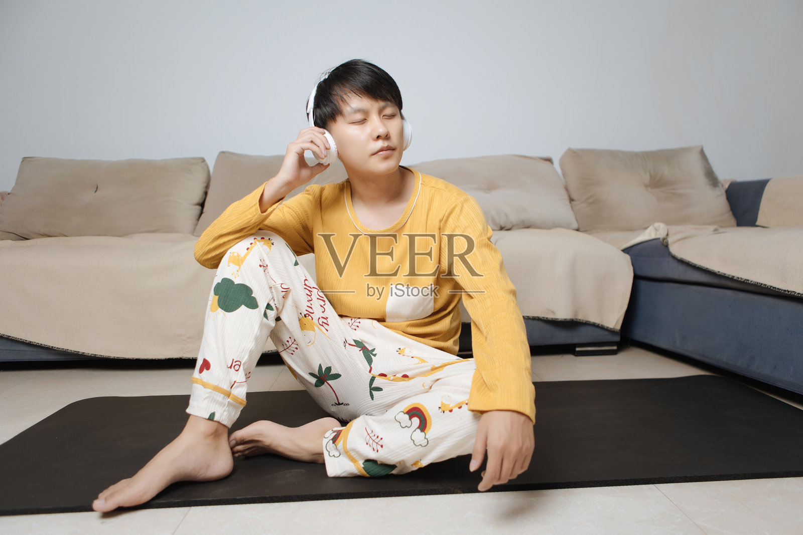 年轻的亚洲男子坐在客厅的瑜伽垫上,戴着耳机,闭着眼睛,聆听音乐,放松身心,注重个人健康、健康生活方式和身体护理,置身于明亮现代的家中。照片摄影图片