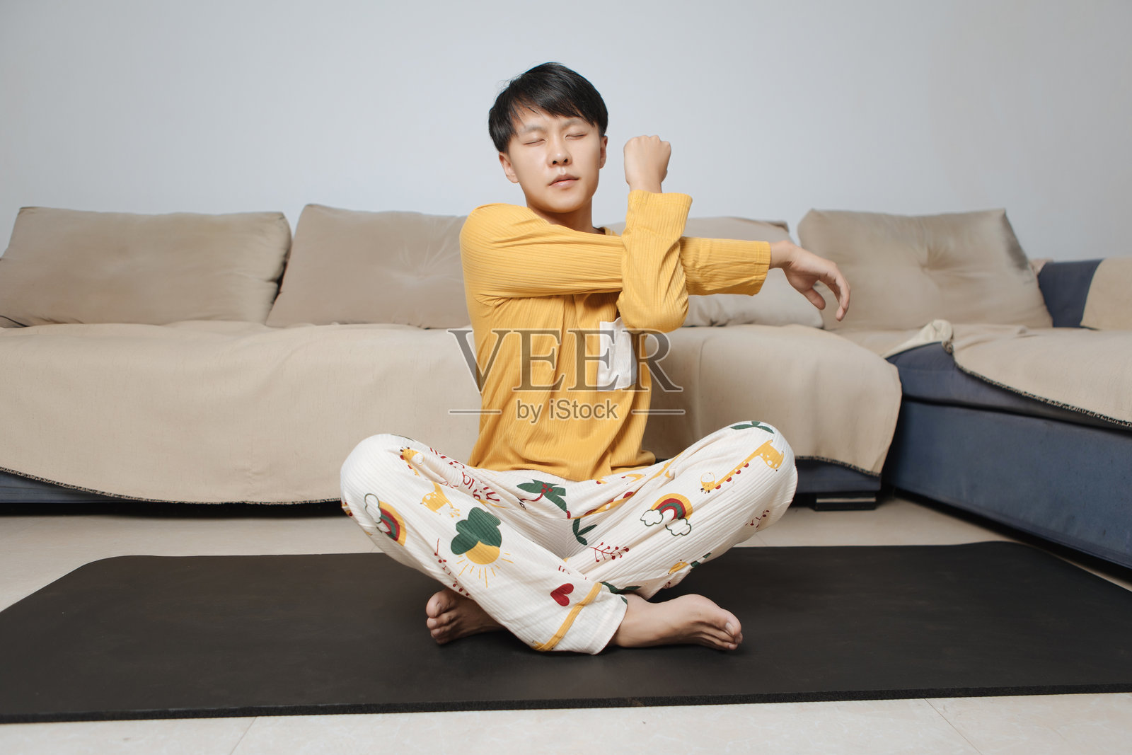 年轻的亚洲男子盘腿坐在客厅的瑜伽垫上,戴着耳机伸展肩关节,践行健康的生活方式和身体护理,专注于明亮现代家居中的个人健康和室内健身。照片摄影图片