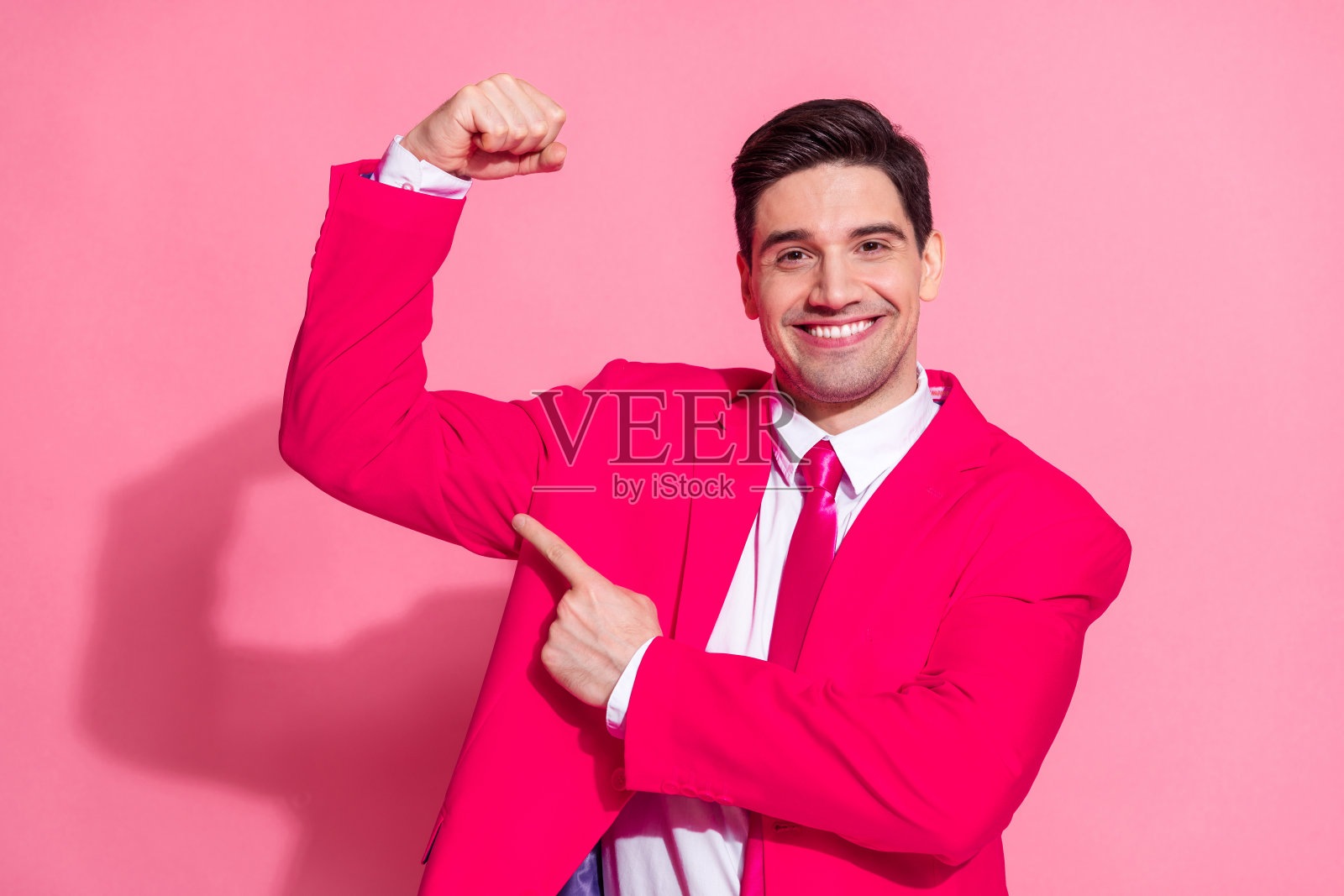 自信迷人的年轻男子,身穿粉色西装,炫耀着手臂肌肉,在充满活力的粉色背景前微笑。照片摄影图片