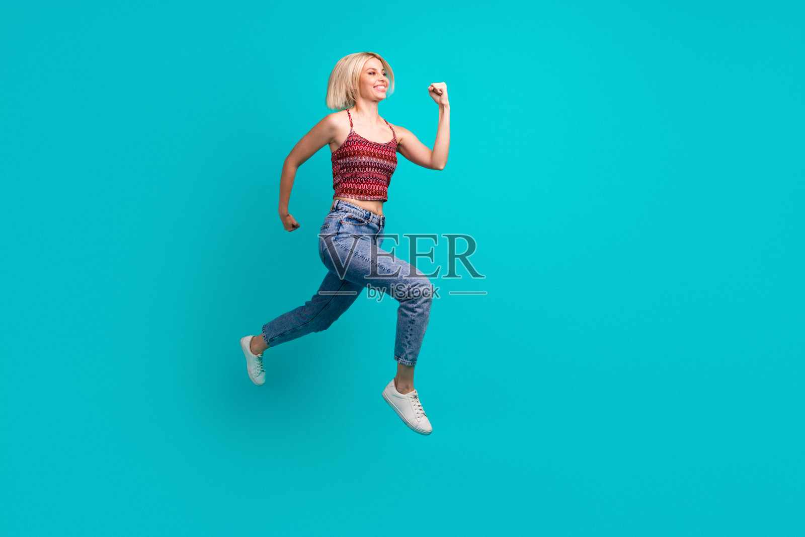 身穿坦克背心、牛仔裤和白色运动鞋的年轻女模特在绿松石色工作室背景下跳跃照片摄影图片