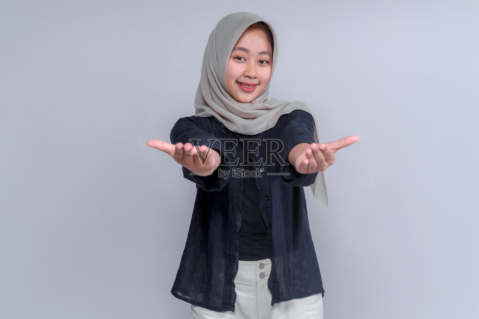 一位年轻的亚洲女性,戴着头巾,身穿黑色衬衫,面带微笑,伸出手做出欢迎的姿势。这张照片传达出友好、温暖和积极的氛围,非常适合商业用途。照片摄影图片
