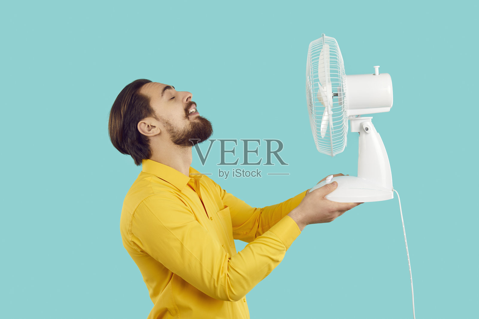 一个留着胡子的男人,在浅蓝色背景下,用电风扇吹风,享受新鲜空气,躲避炎热。照片摄影图片