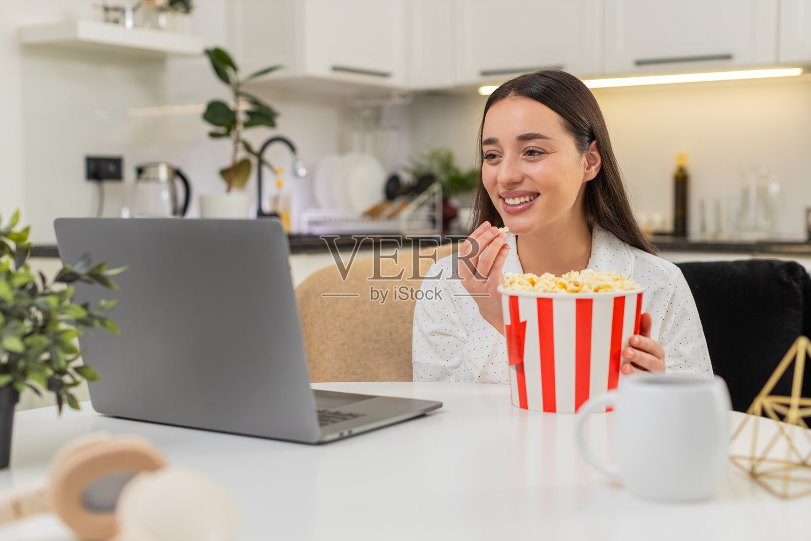 年轻女子在舒适的夜晚享受拖车视频的乐趣,她大笑着,旁边放着爆米花和笔记本电脑,观看流媒体视频。照片摄影图片