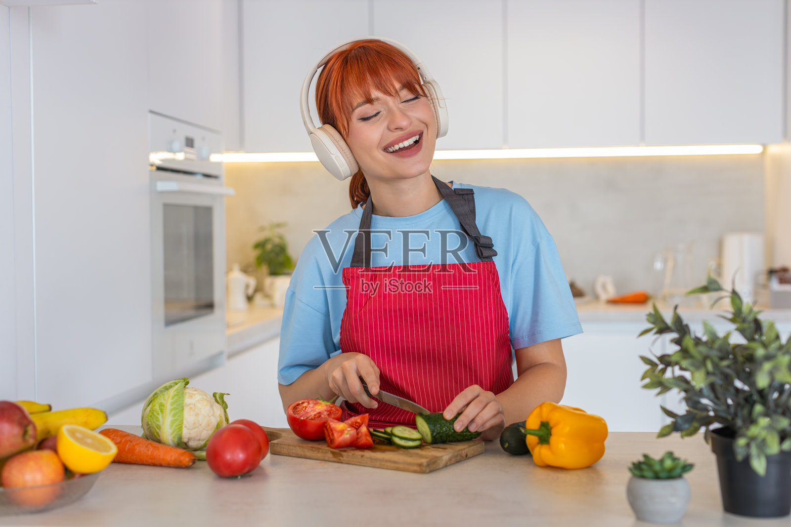 年轻女子在砧板上切黄瓜和番茄,制作色彩缤纷的沙拉,并品尝一口新鲜的美味,脸上带着微笑照片摄影图片