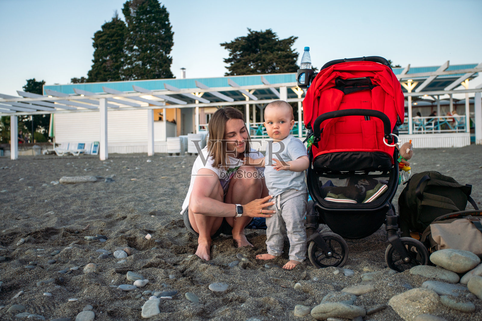 妈妈带着蹒跚学步的孩子和红色婴儿车在海滩上,带着装备和水瓶的家庭外出游玩照片摄影图片