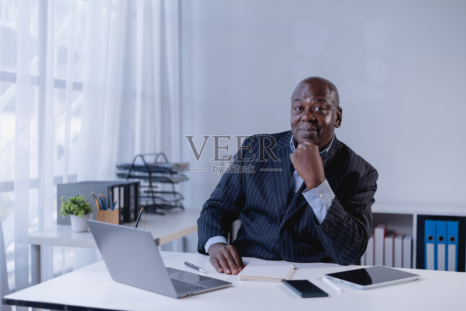 一位穿着细条纹西装的年长非裔美国商人,自信地坐在办公室书桌前,桌上有笔记本电脑、平板电脑和智能手机,他正直接看向镜头。照片摄影图片