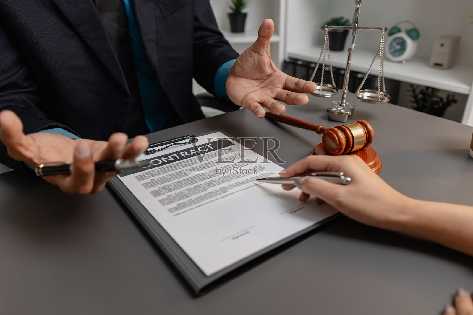 一位身穿西装的男律师正在与一位女客户讨论合同细节,并指向法律文件上的签名处。办公桌上放着象征法律与正义的法槌。照片摄影图片