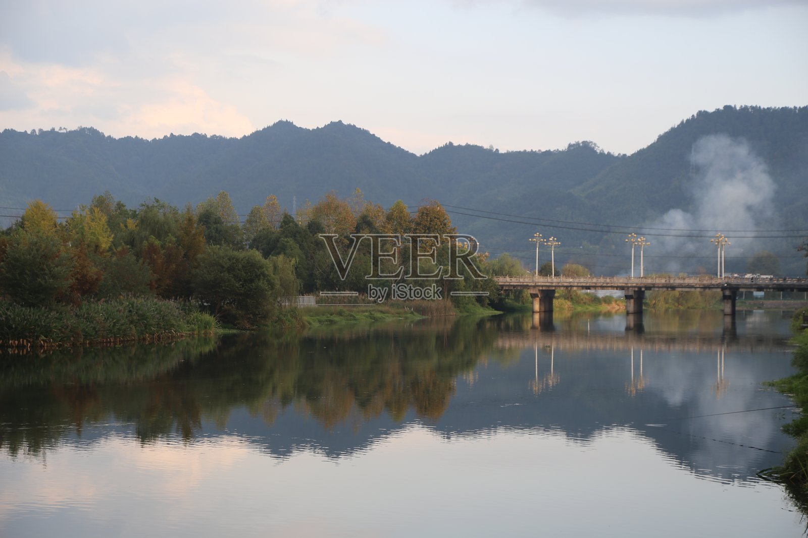 秋日傍晚,河水两岸绿树成荫,倒映着远处的山峦与云彩。照片摄影图片