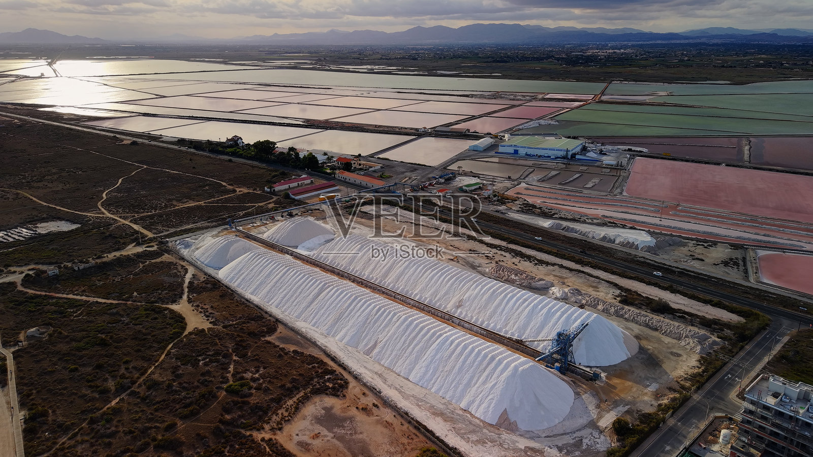 西班牙圣波拉附近,无人机视角拍摄的大型白色盐堆和色彩缤纷的蒸发池,傍晚时分,远山与水面倒影清晰可见。照片摄影图片