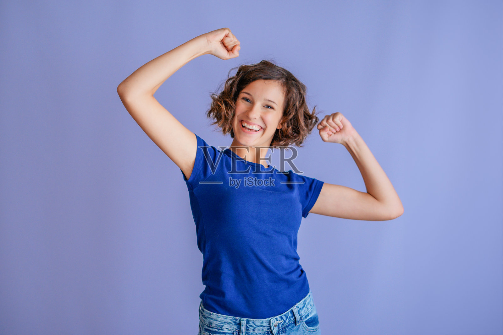 一名快乐的女学生在充满活力的蓝色工作室背景前,举着拳头,脸上带着喜悦的表情,庆祝学业成功。照片摄影图片