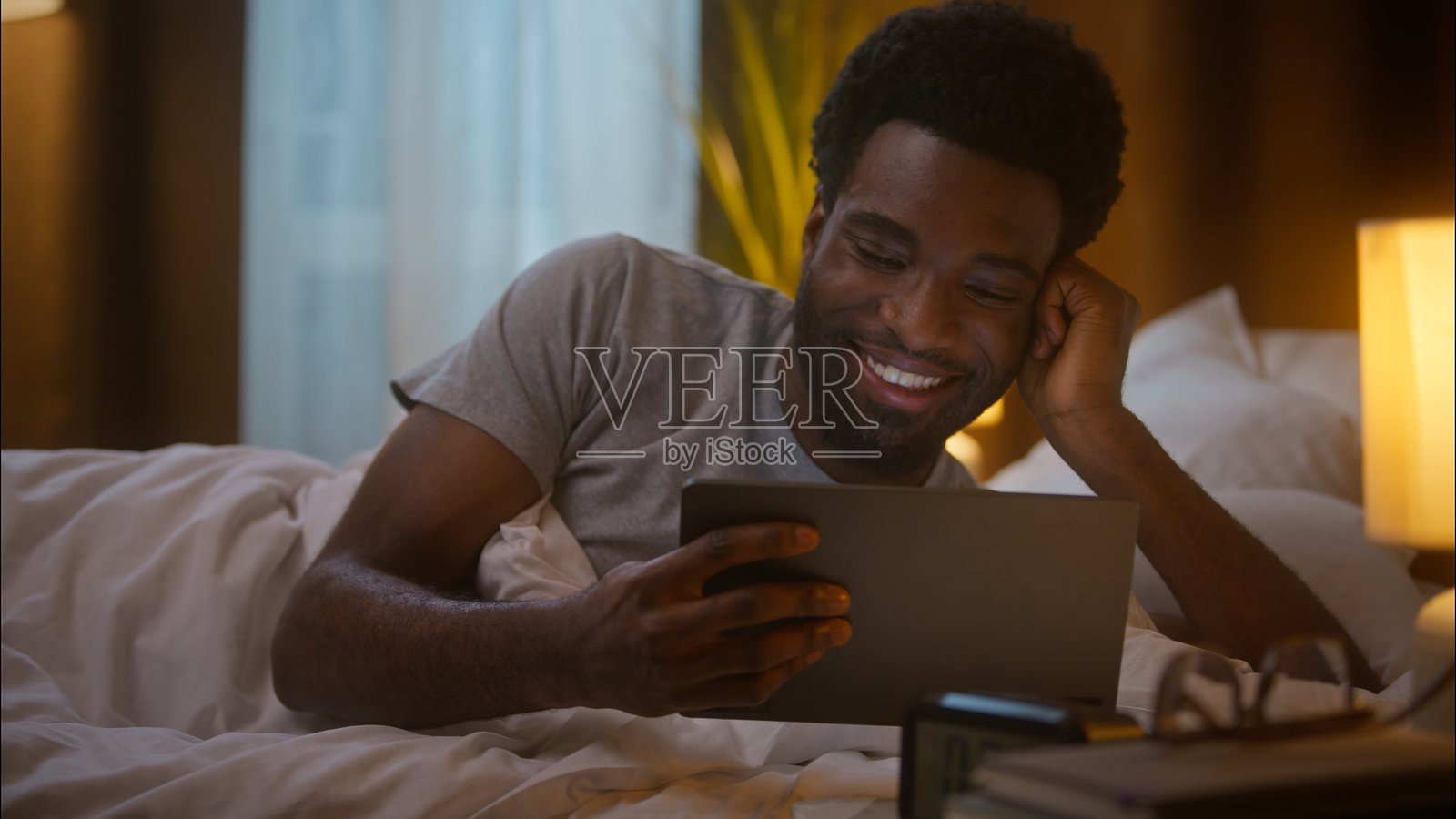 快乐大笑的非裔美国人,客户、自由职业者,在家中卧室使用数字平板电脑观看连续剧、搞笑视频、社交媒体内容、阅读新闻,触摸屏幕,放松,玩乐。照片摄影图片