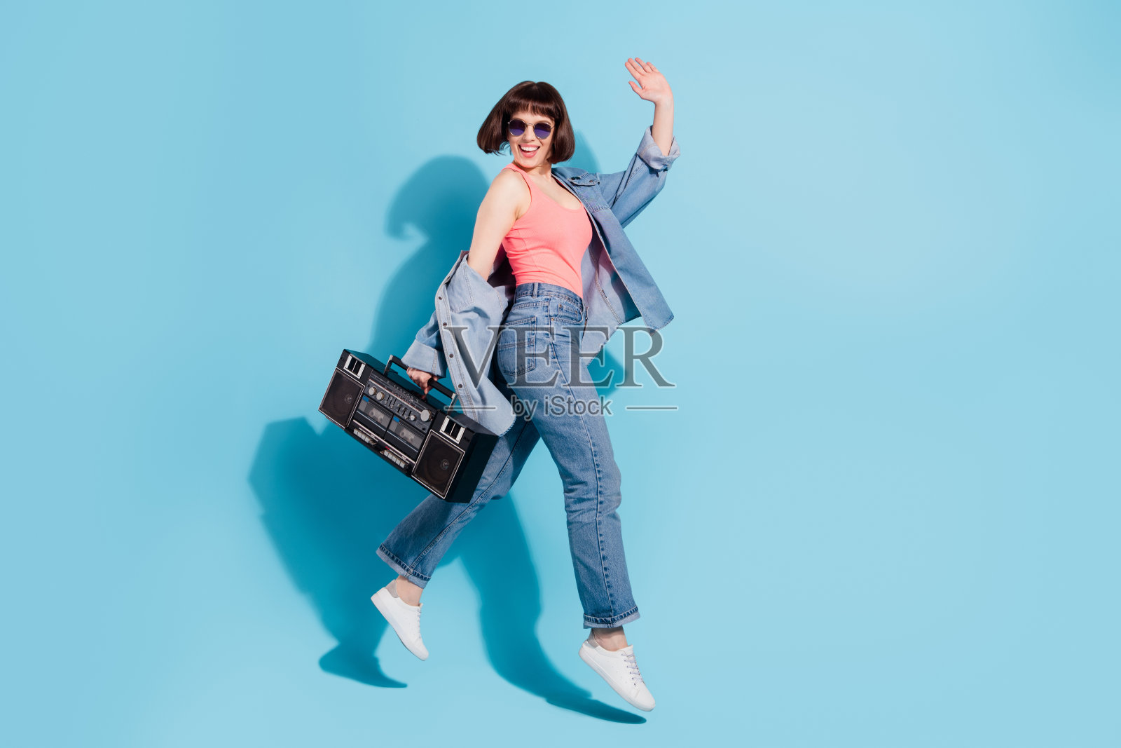 一位漂亮的、快乐的、无忧无虑的女孩,身穿全长服装,手提录音机,在明亮的蓝色背景上跳跃,占据整个画面照片摄影图片