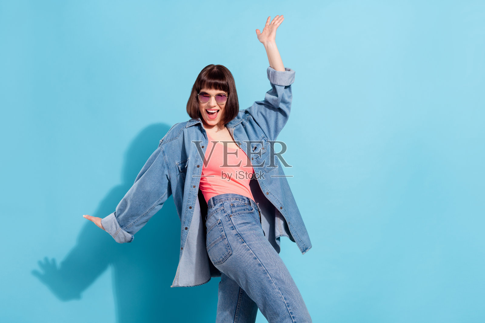 穿着牛仔衬衫、戴着深色眼镜的时尚俏丽年轻女子,在蓝色背景前载歌载舞,笑容灿烂。照片摄影图片