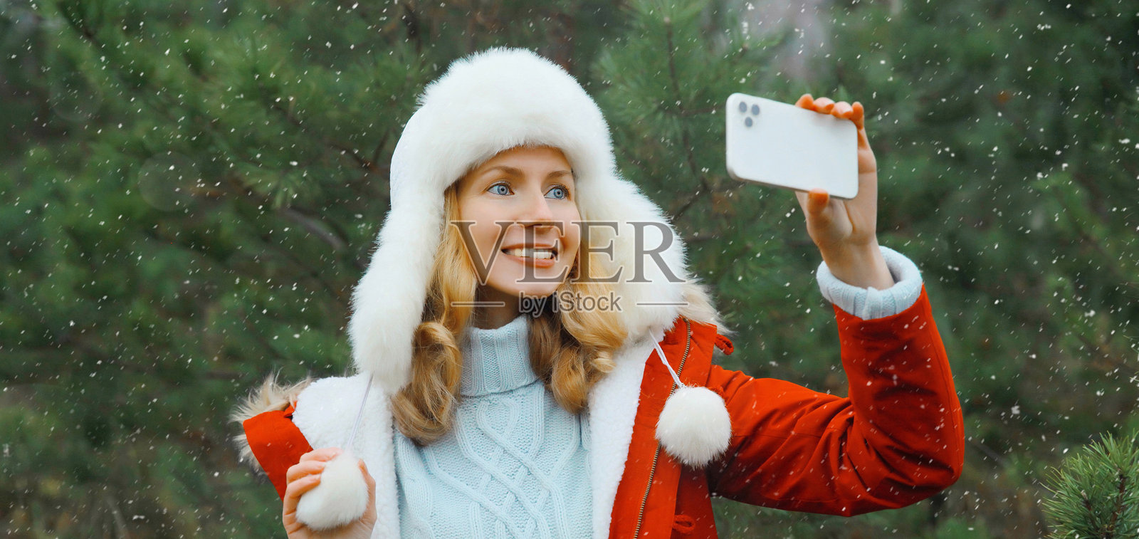 快乐微笑的女人在冬日公园里,以圣诞树为背景,用智能手机自拍照片摄影图片