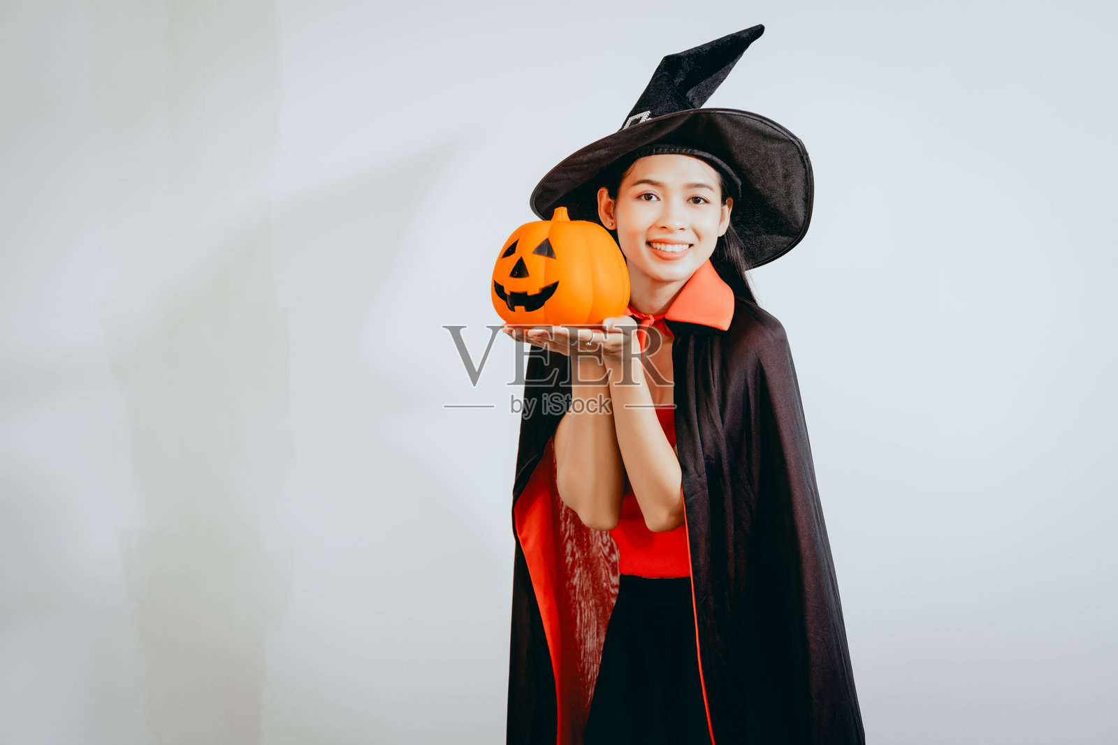 一位打扮成女巫的美丽亚洲年轻女子,手持南瓜,在干净的白色背景前欢庆万圣节,散发着节日的精神、幸福和俏皮的节日魅力。照片摄影图片