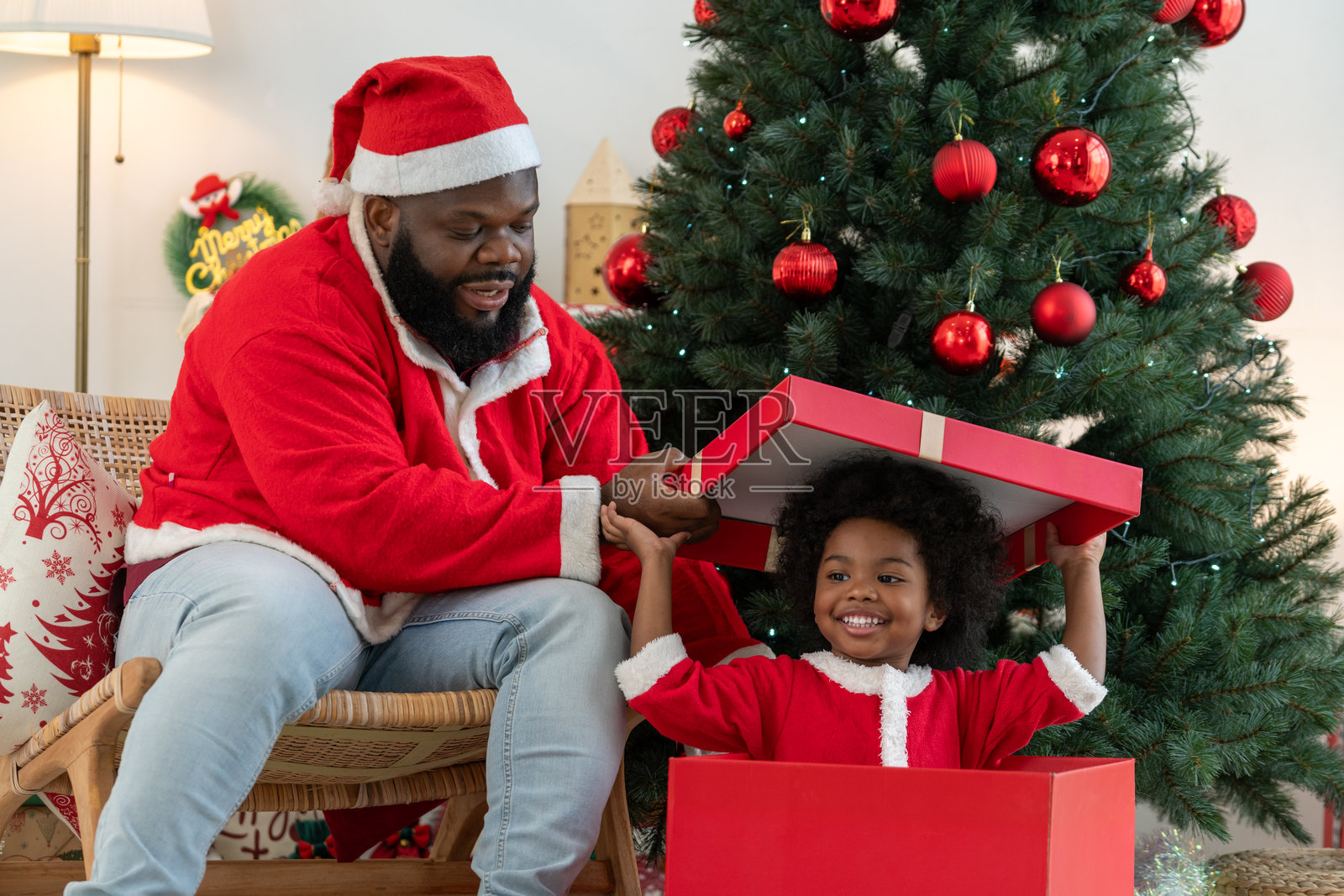 快乐的黑人小男孩和穿着圣诞老人服装的爸爸坐在巨大的圣诞礼物盒里,一家人正在庆祝圣诞节假期。照片摄影图片