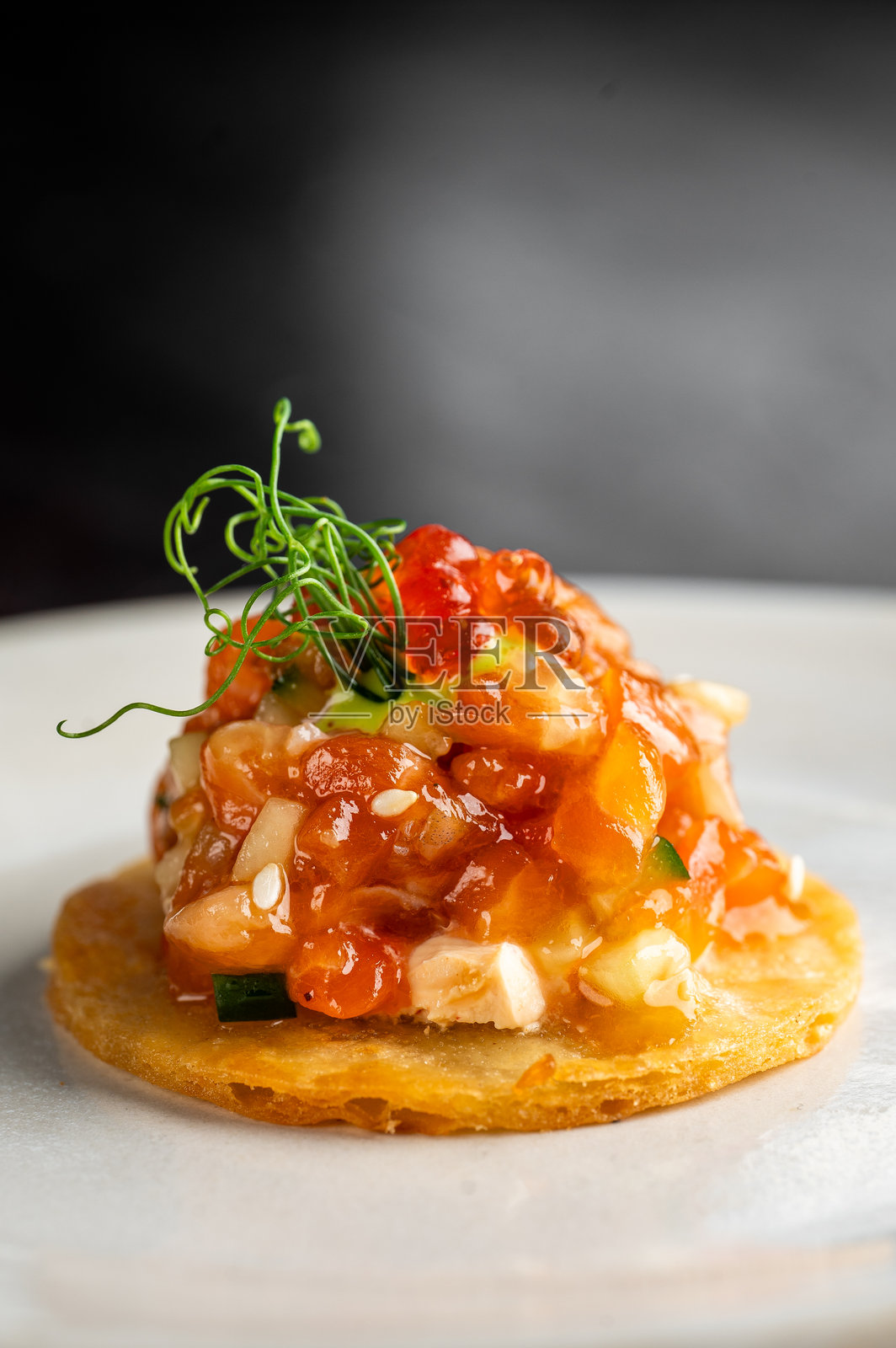 酥脆的金黄色玉米饼上,摆放着新鲜的三文鱼鞑靼开胃菜,上面点缀着三文鱼籽和微型蔬菜。这道菜盛放在一个白盘中,背景为深色。照片摄影图片